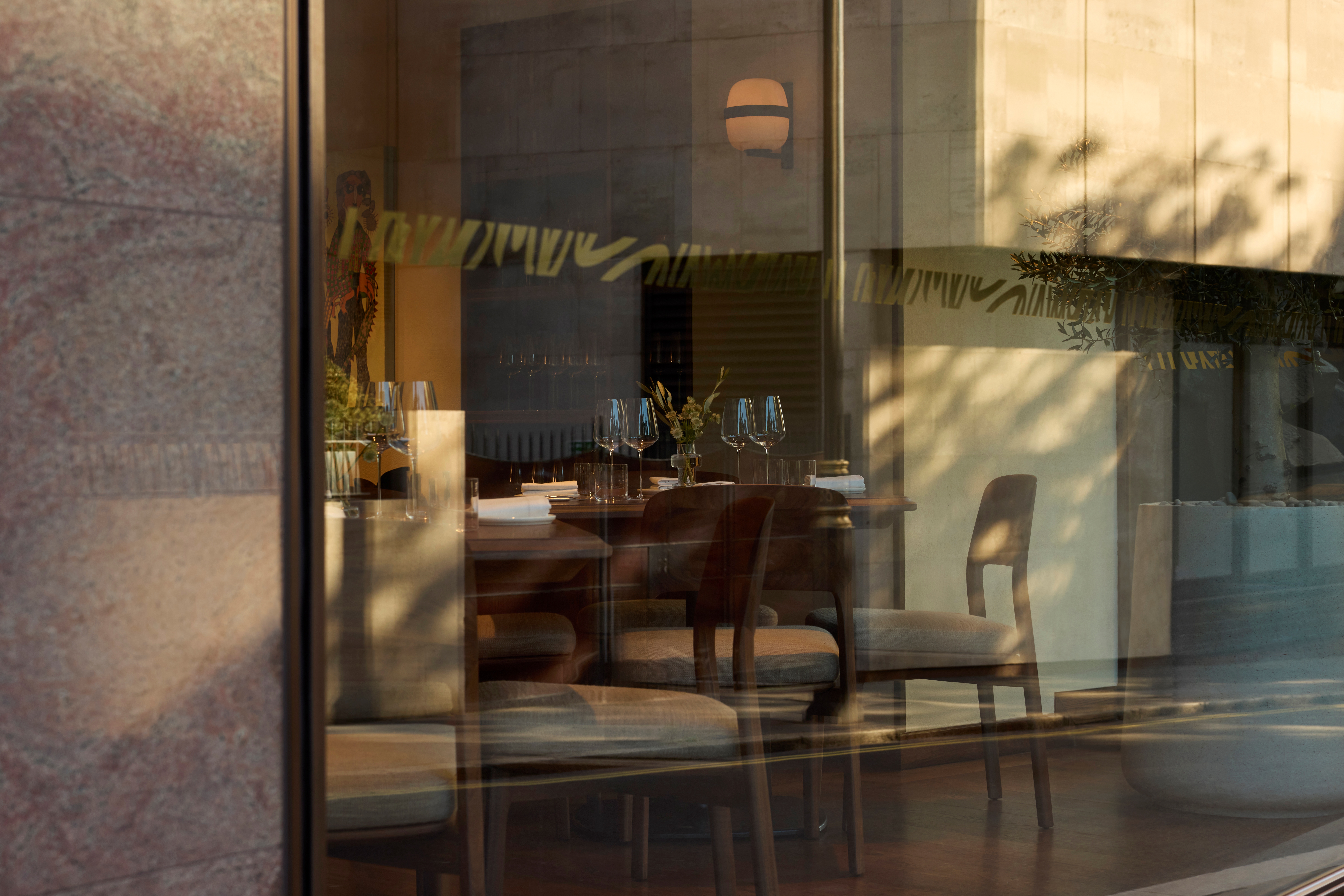 A beautifully and naturally lit glass facade of a restaurant projects a warm glow onto the front of the eatery while letting passersby peek inside its wood-clad, sophisticated interiors.