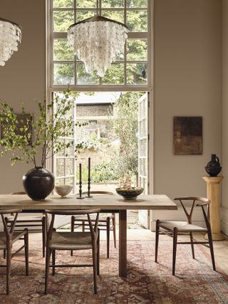 A warm beige-gray dining room with high ceilings and boho-inspired pendant lights hanging. There is a wooden dining table with wooden chairs around it and a black vase that has green stems in it. The back wall has open french doors.