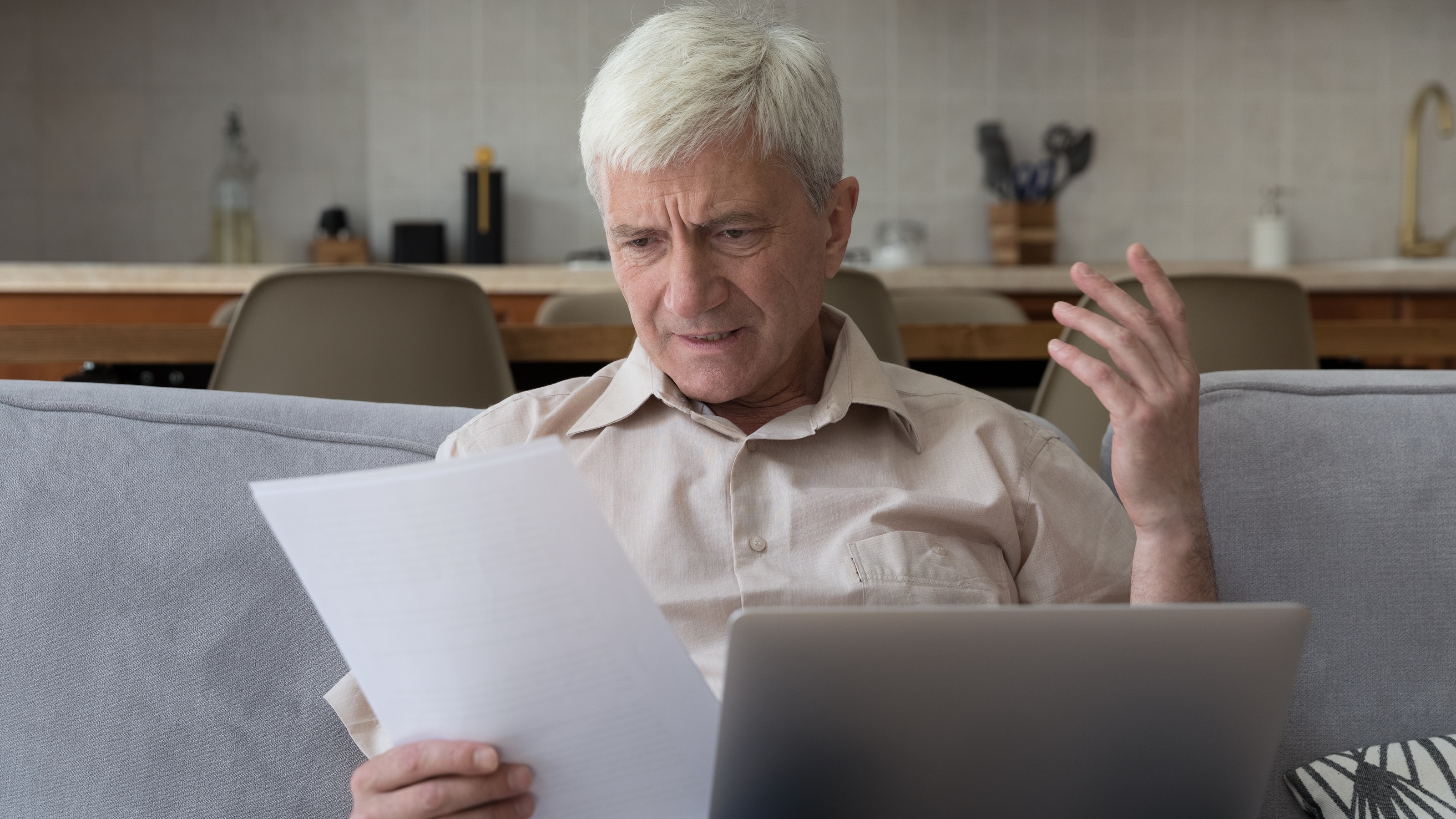 An older man looks a bit confused as he looks at paperwork on his sofa with his laptop.