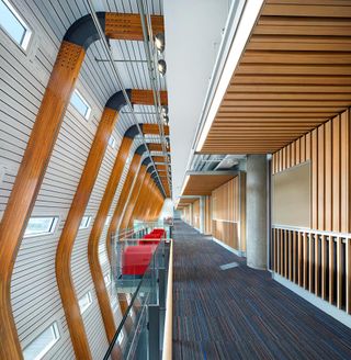 A hallway in Vancouver’s University of British Columbia. There is a carpet on the floor, with concrete columns supporting the upper level. Wooden beams serve as decoration, with wooden boards covering the walls.