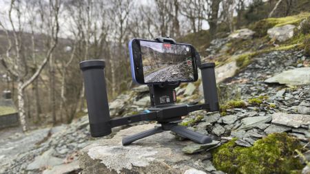 Smartphone mounted in a dual-handle rig with mini tripod feet recording video on a rocky hillside.