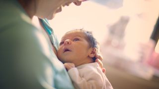 A mom is shown smiling at her newborn baby who she is holding against her chest. The baby is looking up at her face. The background is blurred.