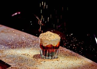 a fluted rocks glass holds a white Russian cocktail. heaps of tahini whipped cream crown the drink. a flurry of sesame seeds rains on the cocktail from above