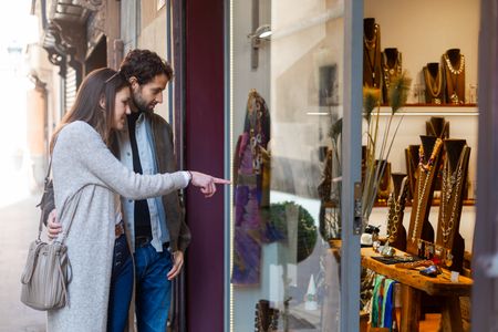 Young couple smiling and pointing at beautiful jewelry displayed in a shop window, sharing a moment of joy and desire for luxury items