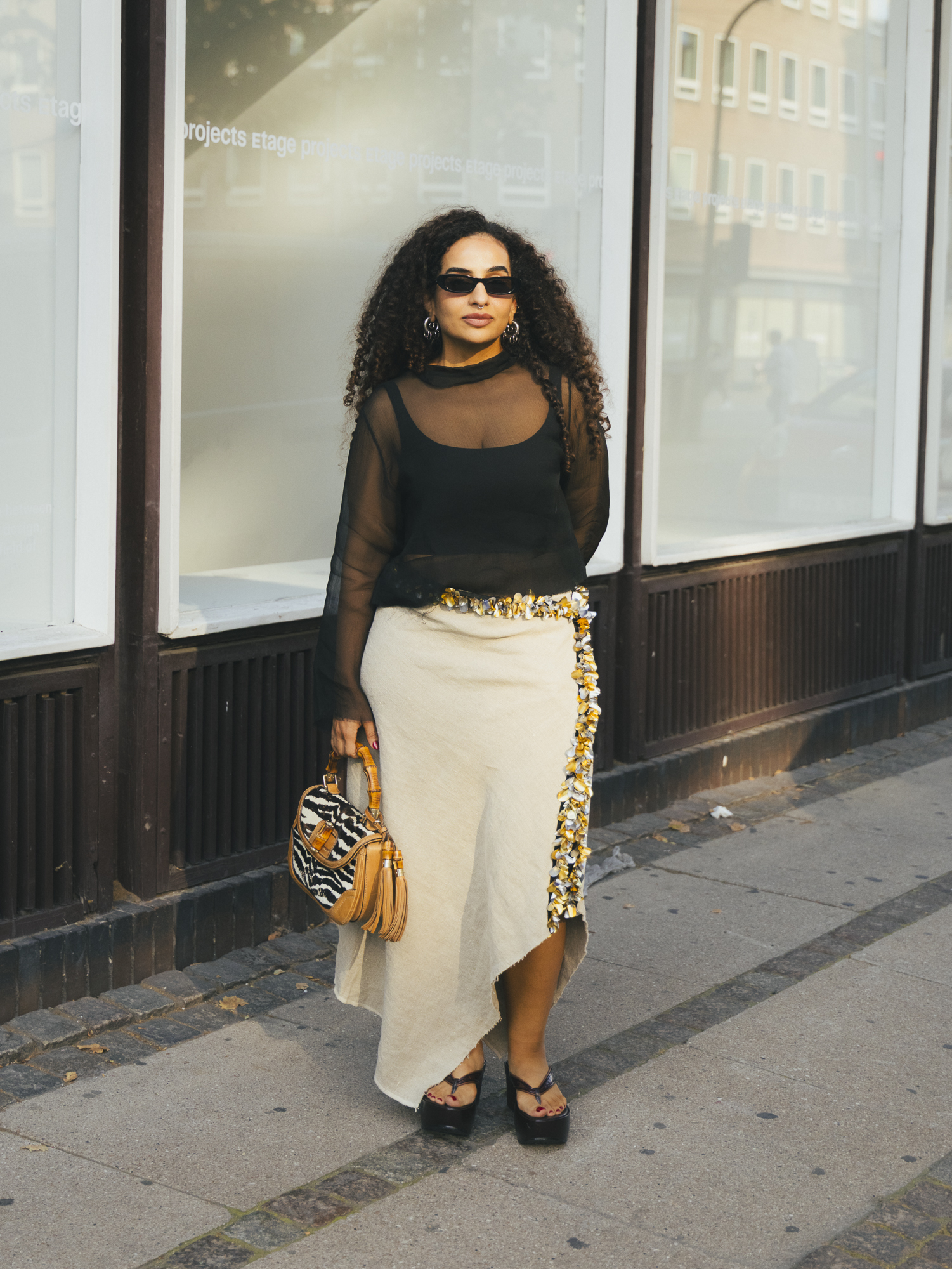 a woman wears a black sheer top, beige skirt, sequin belt, and platform flip-flops.