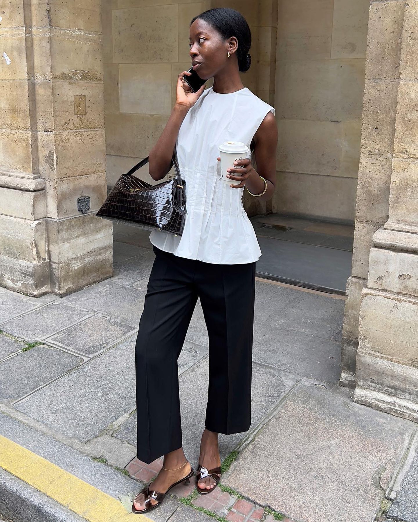 influencer and stylish Sylvie Mus poses while on her phone and holding a coffee, wearing a white sleeveless pleated top, a brown croc-embossed bag, cropped black pants, and brown patent mule kitten-heel sandals