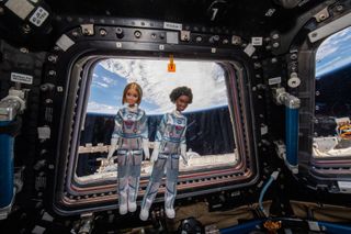 Two dolls, one with dark skin and one with light skin, are wearing silver astronaut suits float in front of a window in the cupola of the International Space Station. Part of Earth can be seen through the window.