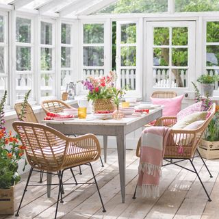 conservatory with white woodwork, rustic table, rattan chairs with metal legs, pretty spring table setting with flowers in basket on table, pink blanket, flowers in crates, pink cushions, wooden floor boards