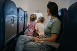two kids on a flight looking out of the aeroplane window, while mum looks on from her seat