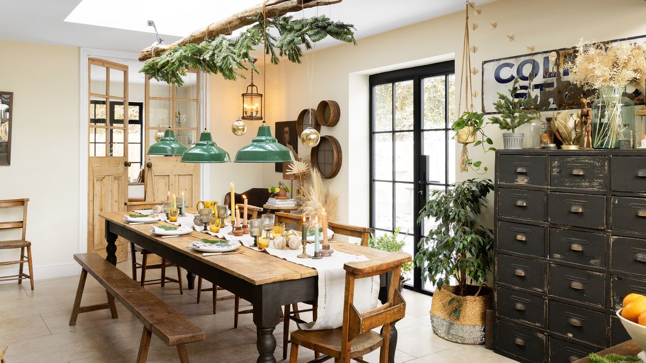 dining room with patio doors with long wooden dining table, antique school chairs and bench with a branch hanging overhead covered in greenery with three green pendant lights