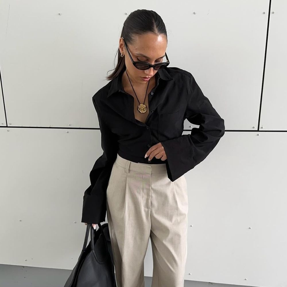 Influencer wears beige trousers tucked into a black blouse. She stand outside and holds a large black handbag and styles it with brown shoes and a gold cord necklace.