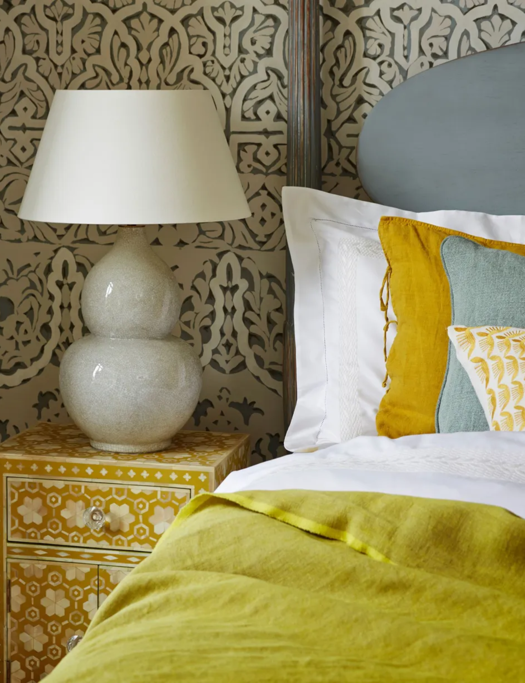 A bedroom with patterned wallpaper, a yellow patterned bedside table with a grey ceramic lamp with white lampshade, and large bed with duck egg blue headboard with bedpost, white bedding, a chartreuse throw, and yellow and blue cushions