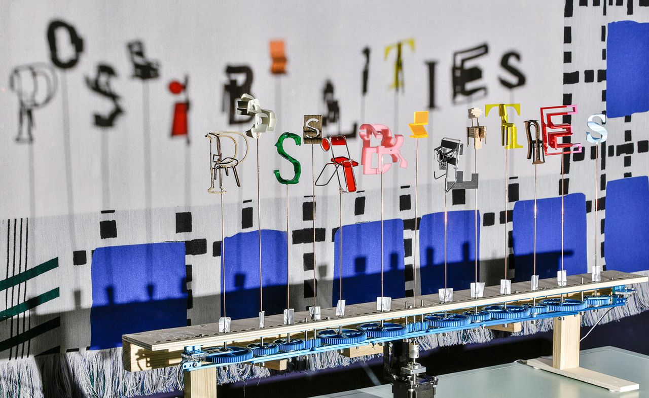 Unique installation gallery piece, neutral metal platform with colourful letters on sticks connected to blue wheel cogs casting shadows behind, white background with blue and black square pattern 