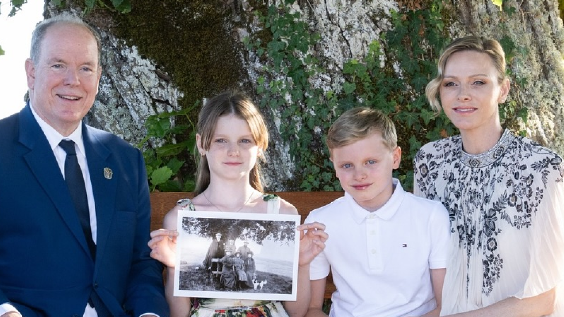 Prince Albert, Princess Charlene and their twins sitting in front of a tree holding a black and white photo