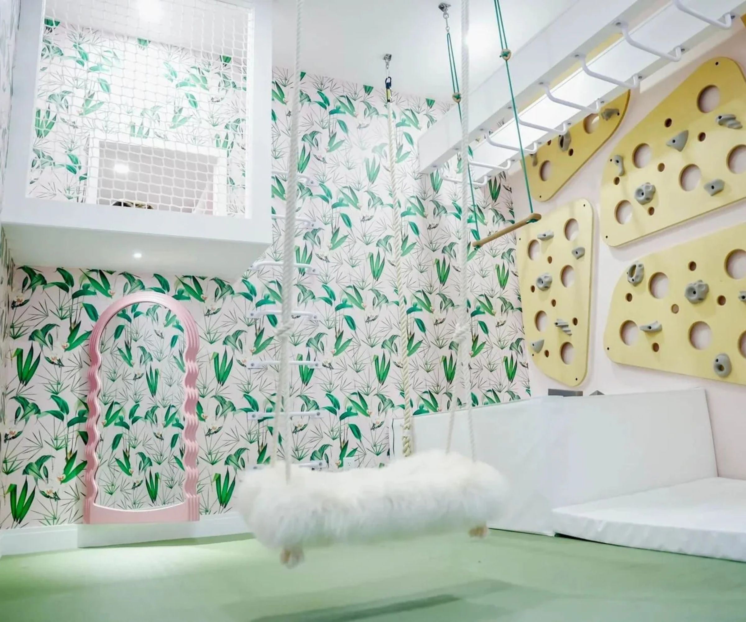 pink and green foliage style wallpapered playroom with climbing frames, a fluffy white faux shearling swing and a pink wavy mirror