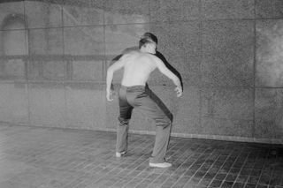 Man wearing no shirt and jeans facing a wall standing in a gorilla like pose.
