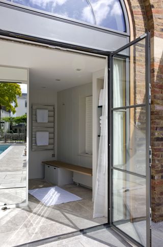 The inside of the pool house has tiled flooring matching that outside, a bench with storage, a mirror, and glass doors