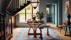 Round table in a blue hallway topped with books and a vase of flowers, sitting on top of a red and blue rug with two small bar stools