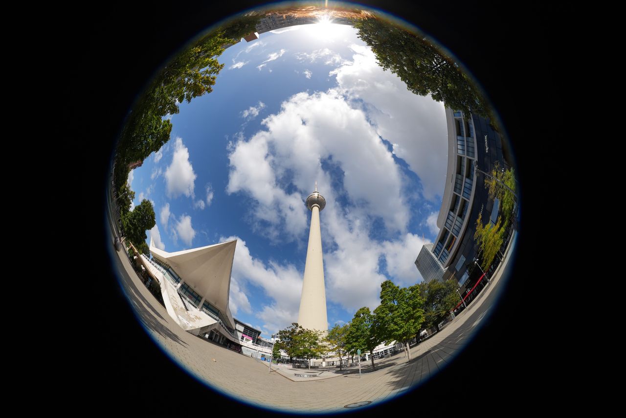 Circular fisheye view looking up at Berlin&rsquo;s Fernsehturm TV tower, surrounded by buildings, trees and a bright blue sky.