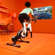 Cyclist riding a Zwift bike with the Zwift app on the wall in front of them in an orange room