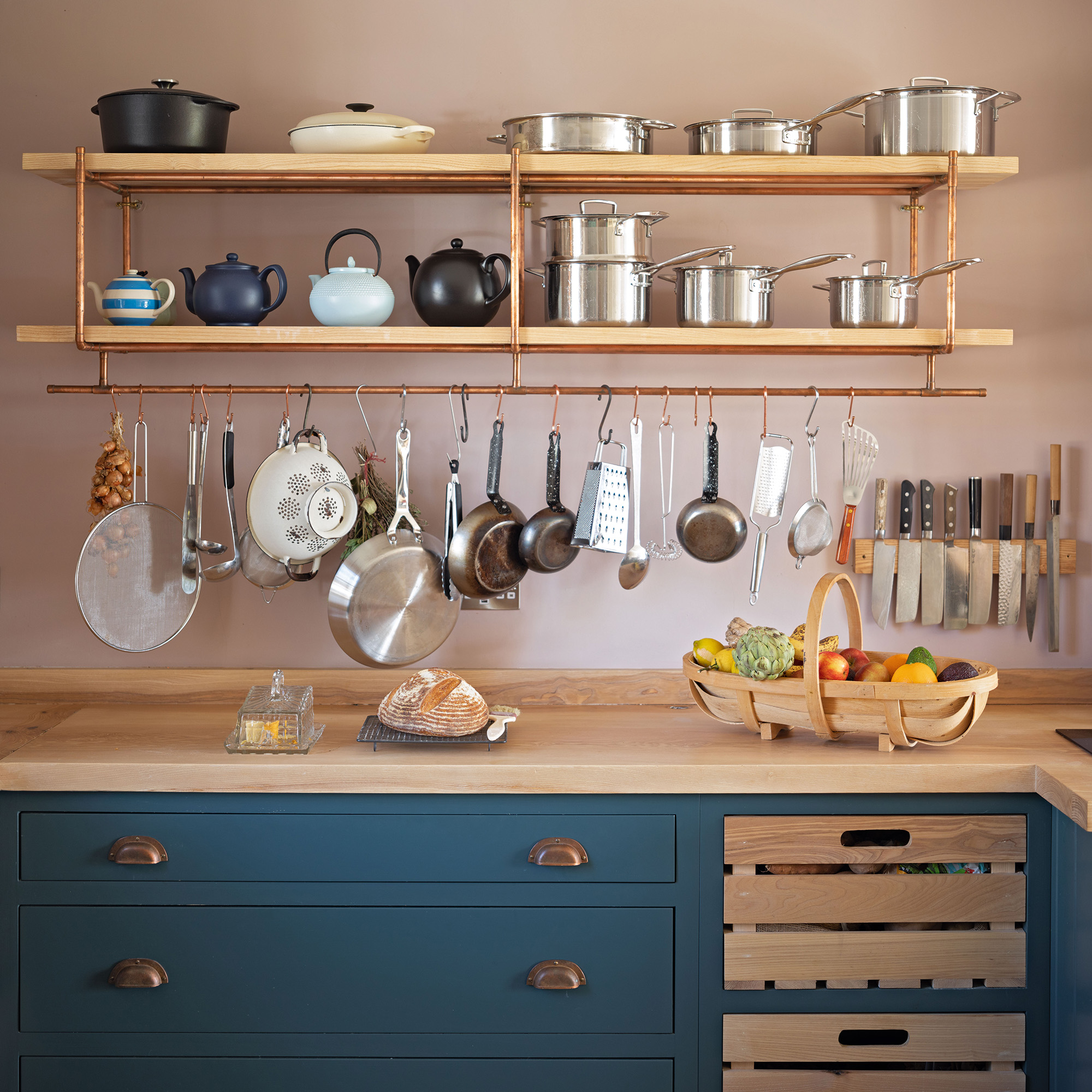 country kitchen with pink wall and blue cabinetry and copper and ash open shelves and hanging kitchen tools