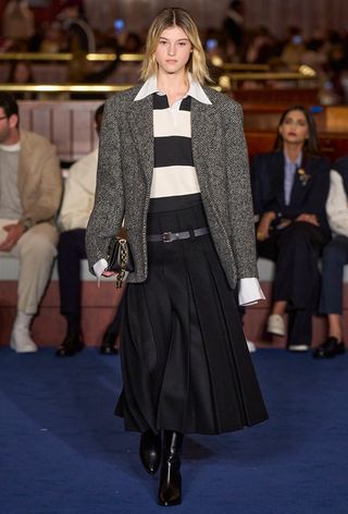 The polo shirt trend on Tommy Hilfiger's spring runway with a model wearing a chevron blazer over a striped polo shirt with a black pleated midi skirt and black boots.