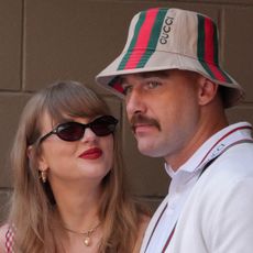 Taylor Swift wears a red gingham dress and black sunglasses and smiles up at boyfriend Travis Kelce, who is wearing a white polo shirt and Gucci bucket hat