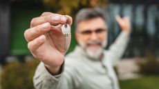 An older man holds out a house key while gesturing to the home behind him.