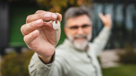 An older man holds out a house key while gesturing to the home behind him.