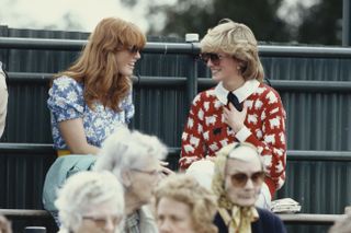 Sarah Ferguson wearing a blue floral dress and smiling, sitting on bleachers next to Princess Diana, who is wearing a red sheep sweater and sunglasses