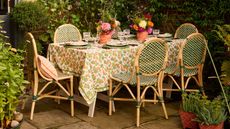 garden party table styled on a patio surrounded by plants with green rattan bistro style chairs, a floral print tablecloth, and potted flower arrangements on the table