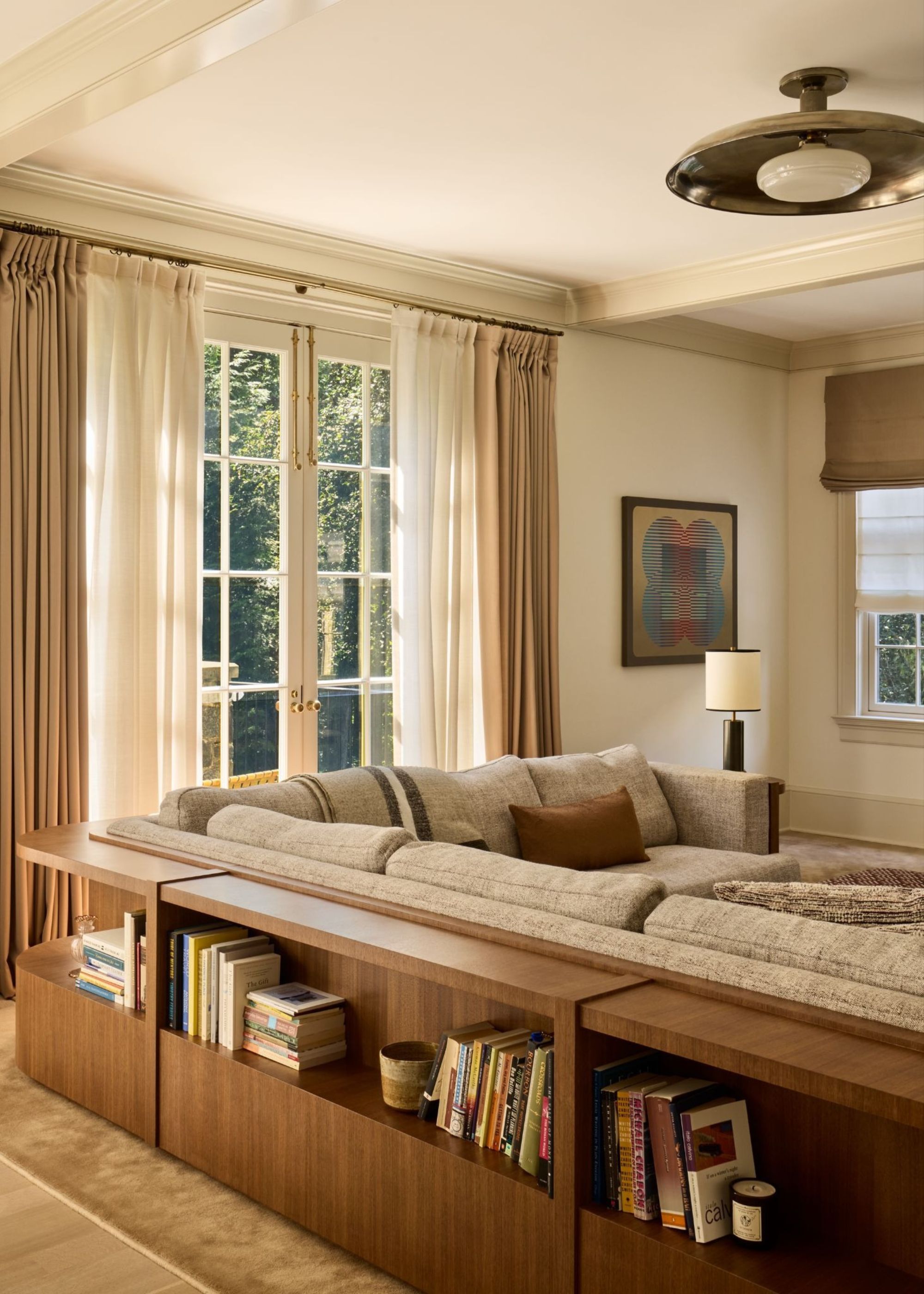 A modern neutral living room with cream walls, French doors with layered neutral curtains, a large taupe corner sofa, and a wooden console table running along the back, with styled books.