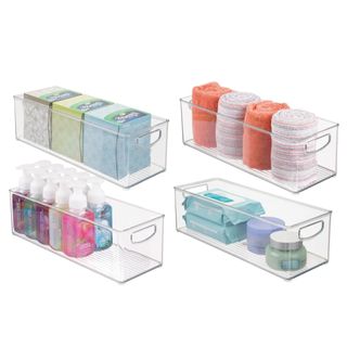 Four clear plastic storage bins with integrated handles and ridged base filled with green, blue and white tissues, orange and striped towels, bottles of handsoap with white lids, and various toiletries