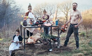Topless male and three children cooking outside on a bench