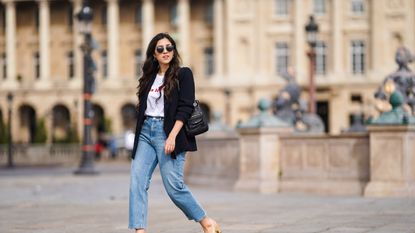 Camille Grandguillote wears sunglasses, a black blazer jacket from Sezane, a black leather bag from Kate Lee, a white t-shirt from "Pas le Time", blue denim jeans pants from Asos, Chanel beige and black slingback shoes, on February 25, 2021 in Paris, France