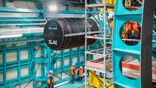 a massive black cylinder is lifted by crane into a round metal enclosure in a large warehouse