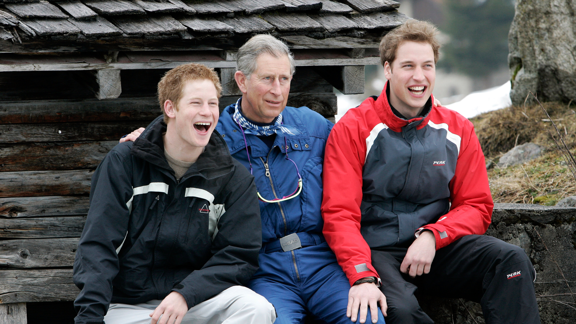 Prince William, Prince Harry and King Charles sitting next to each other wearing ski outfits and laughing