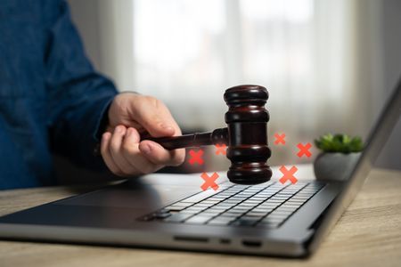 A hand holding a gavel striking a laptop with small red crosses emerging. 