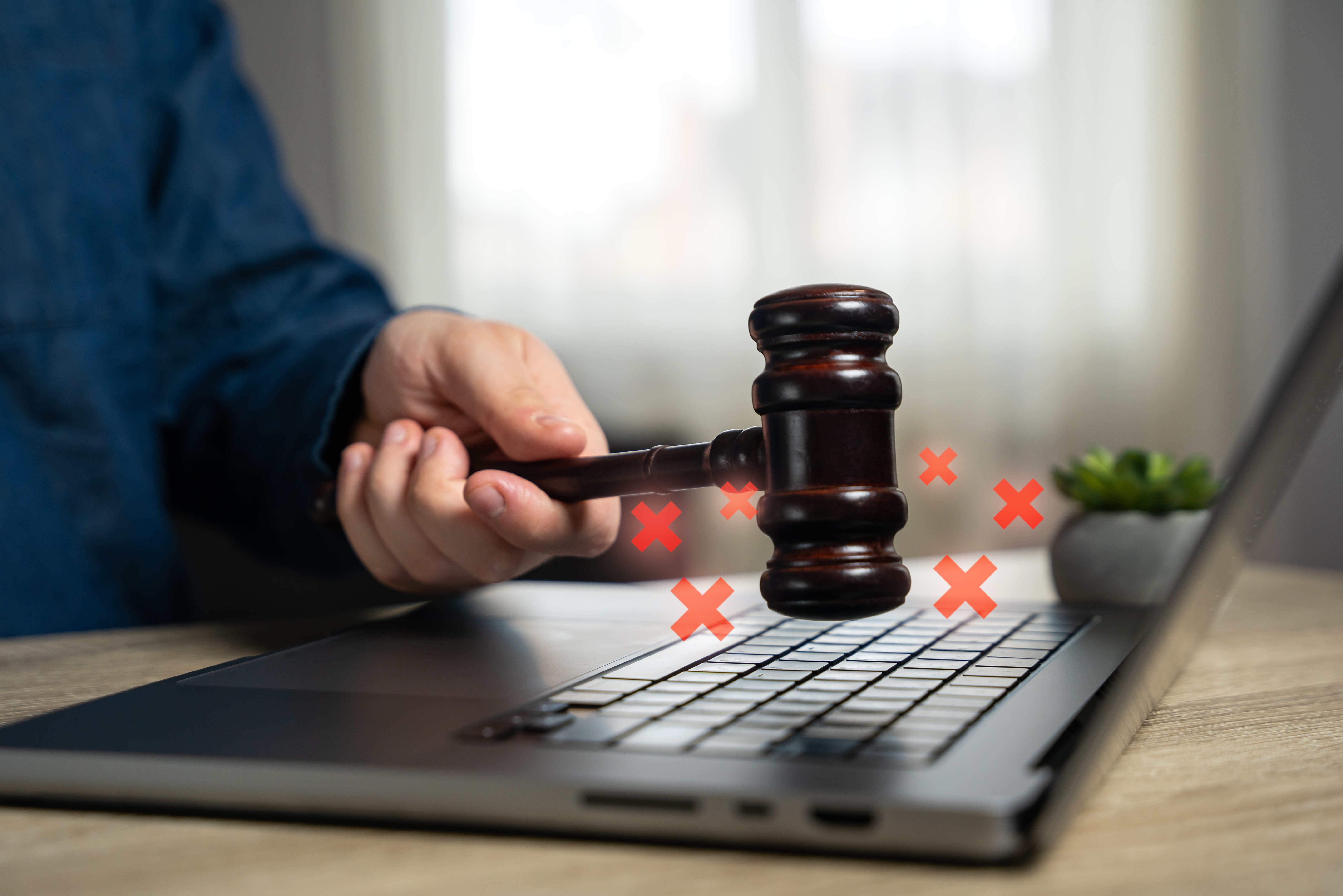 A hand holding a gavel striking a laptop with small red crosses emerging. 