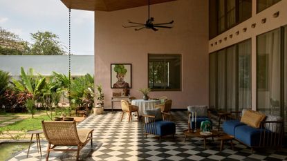 A large patio with outdoor furniture, a sun lounger, an overhead fan, a checkerboard floor, a rain chain, potted plants and framed art