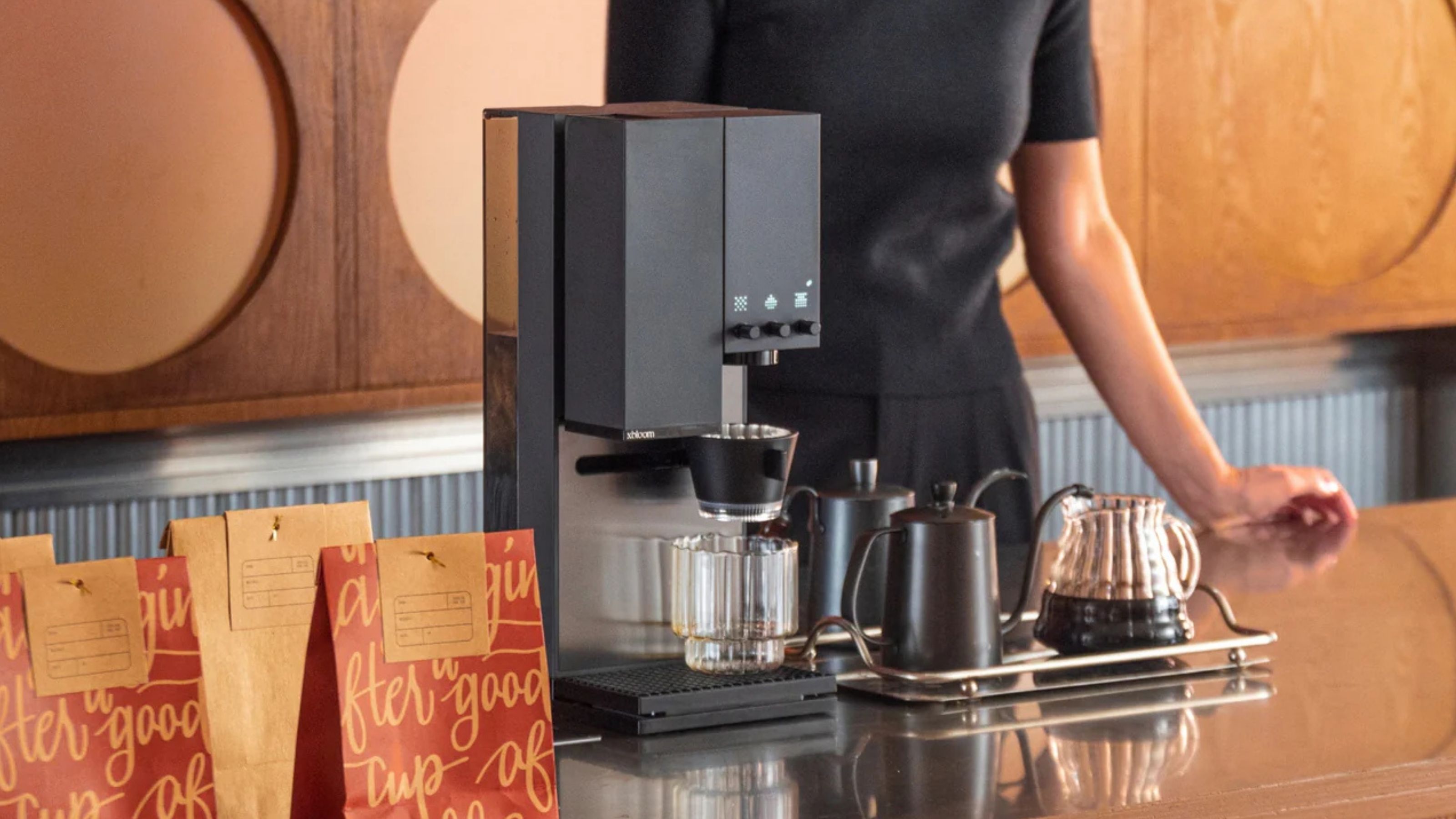Image of a black pour-over coffee machine by xBloom on a reflective countertop in a room with wooden cabinets behind the worktop. There is a person dressed in black behind the coffee machine.