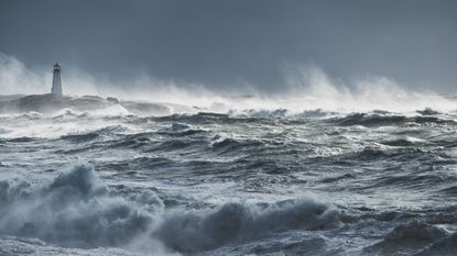 A lighthouse sits in the distance against the backdrop of storm clouds while turbulent waves crash around it.