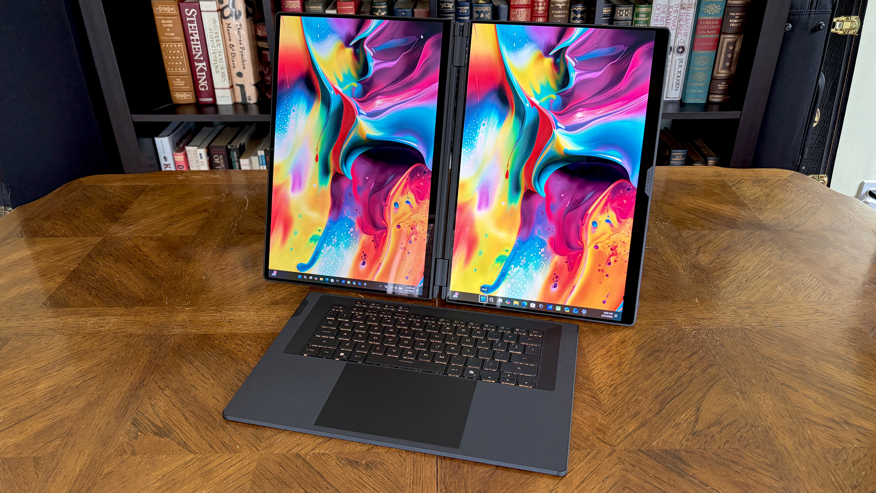 A laptop with dual vertical monitors displaying vibrant abstract art on a wooden table, set against a backdrop of a bookshelf filled with books.