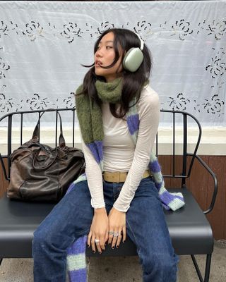 fashion influencer Vivian Li with Apple headphones a colorful striped scarf, long sleeve cream tee, tan suede belt, and dark wash jeans