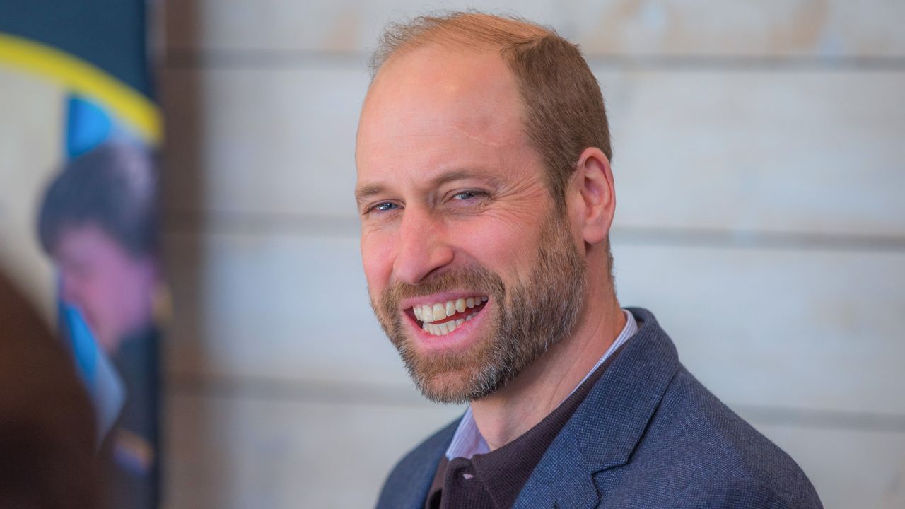 Prince William wearing a blue blazer and laughing in front of a white wall