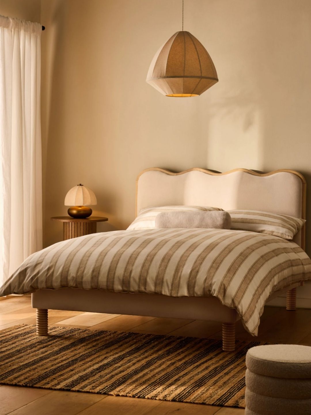 Image of a wooden and upholstered scalloped bed with beige and white striped bedding in a neutral room with a striped rug underneath and a wooden side table that has a lamp on it.