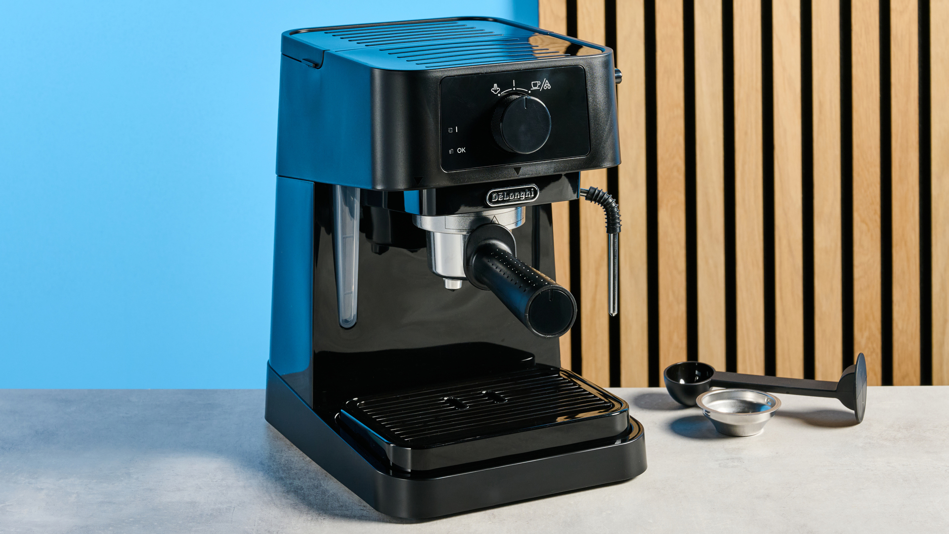 the de&#039;longhi stilosa espresso machine in black, with one portafilter, a dial for espresso and milk steaming, and a plastic tamper, photographed against a blue background