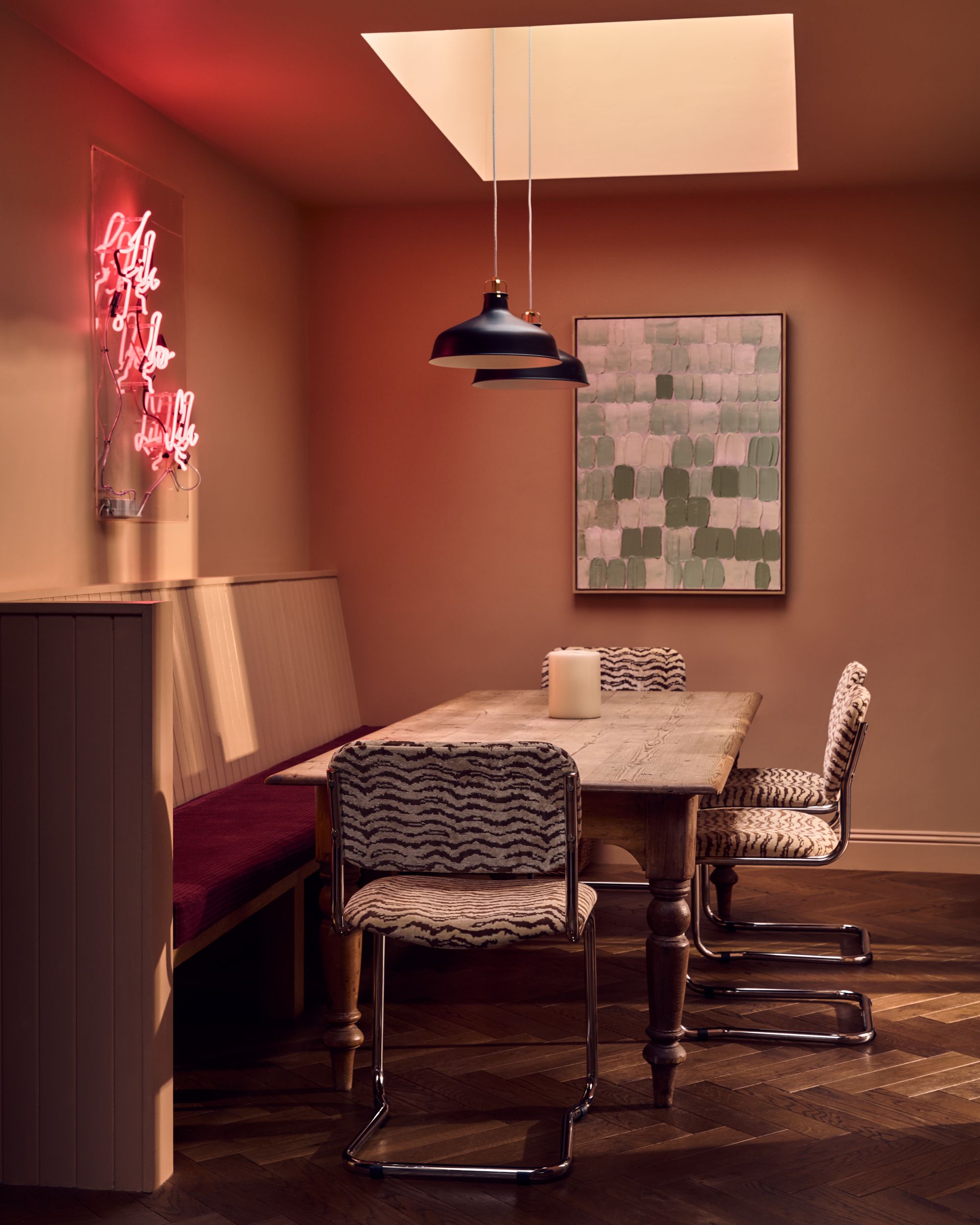 Image of a dusty pink/neutral dining nook area with a built-in bench seat that has a deep red seat, a neon sign on the wall, a rustic table, and four modern, zebra print chairs.