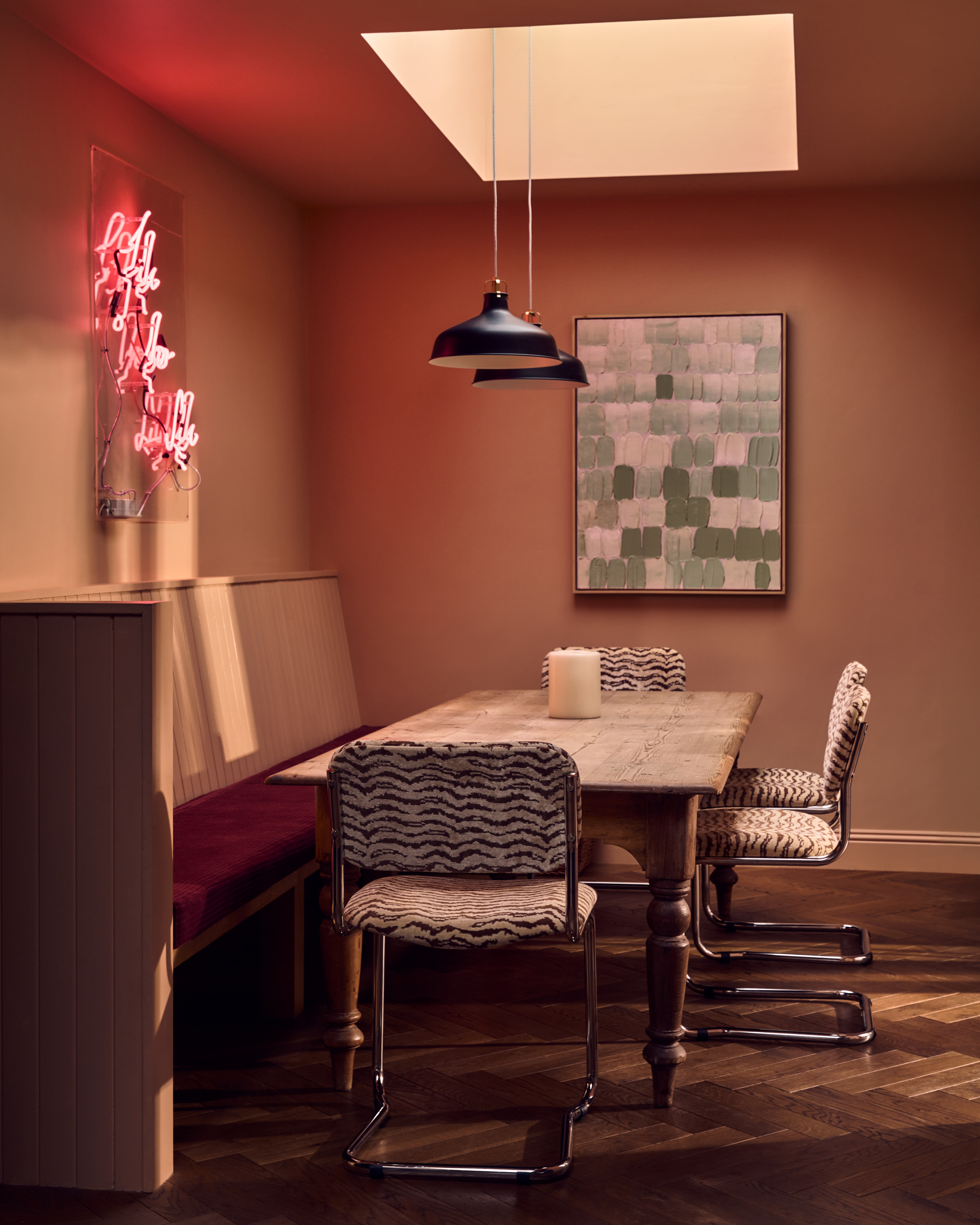 Image of a dusty pink/neutral dining nook area with a built-in bench seat that has a deep red seat, a neon sign on the wall, a rustic table, and four modern, zebra print chairs.