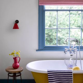 budget bathroom ideas, bold colourful bathroom with blue painted window frame, yellow bath, red vase, red wall light, stripe blue towel, red graphic blind